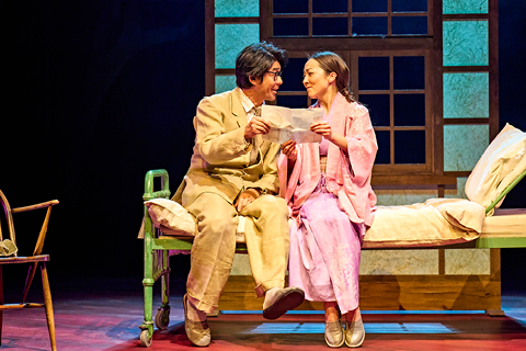 Tatsuo (Dai Tabuchi) and Yasuko (Emily Piggford) sit on a hospital bed. Dai wears a light brown linen suit and Emily wears a pink dress and gown over the top. They hold a letter and smile at each other with affection.