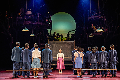Miss Hara (Arina Ii) and members of the My Neighbour Totoro company. Arina wears a pink skirt and white shirt and stands in front of a blackboard. Members of the company face her in lines, as if in a schoolroom.