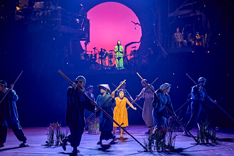 Members of the My Neighbour Totoro company. Satsuki (Ami Okumura Jones) wears an orange dress and stands behind a group of the company, who hold long sticks. They are in shadow. Behind them stands you can see an outline of the orchestra.