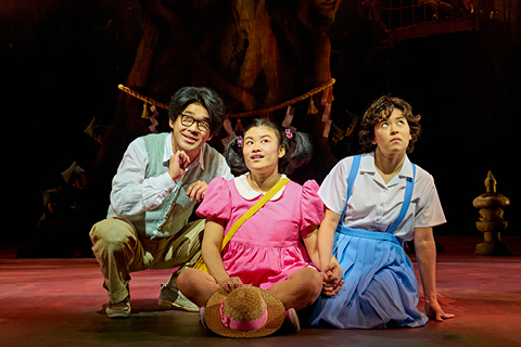 Tatsuo (Dai Tabuchi), Mei (Mei Mac) and Satsuki (Ami Okumura Jones) sit on a stage. Dai wears brown trousers, a white shit and mint green waistcoat. Mei wears a pink dress with a white collar and a yellow cross-body bag. Ami wears a blue pinafore dress and white shirt.