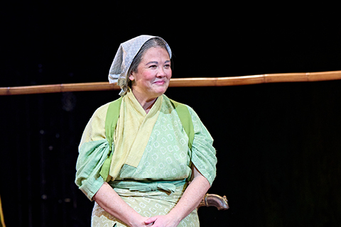 Granny (Jacqueline Tate) stands on a stage, wearing a green tabard and apron and light green trousers, with a grey headscarf.