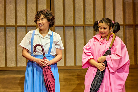 Satsuki (Ami Okumura Jones) holds a folded up red umbrella and wears a blue pinafore dress and white short-sleeved shirt, with blue wellington boots. Mei (Mei Mac) stands nearby, wearing a long pink rain shawl and pink wellington boots, holding a black umbrella.